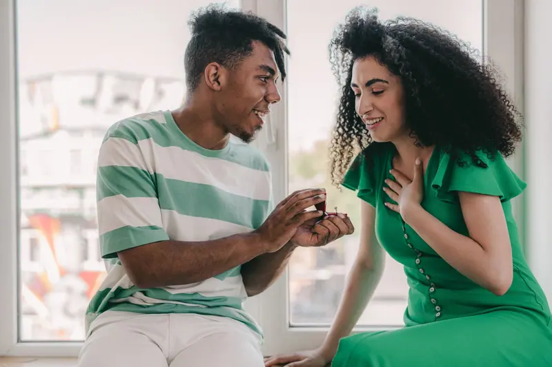 A man presenting an engagement ring to a smiling woman by a bright window