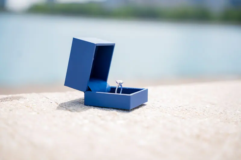 Engagement ring in an open blue box on a stone ledge by the water